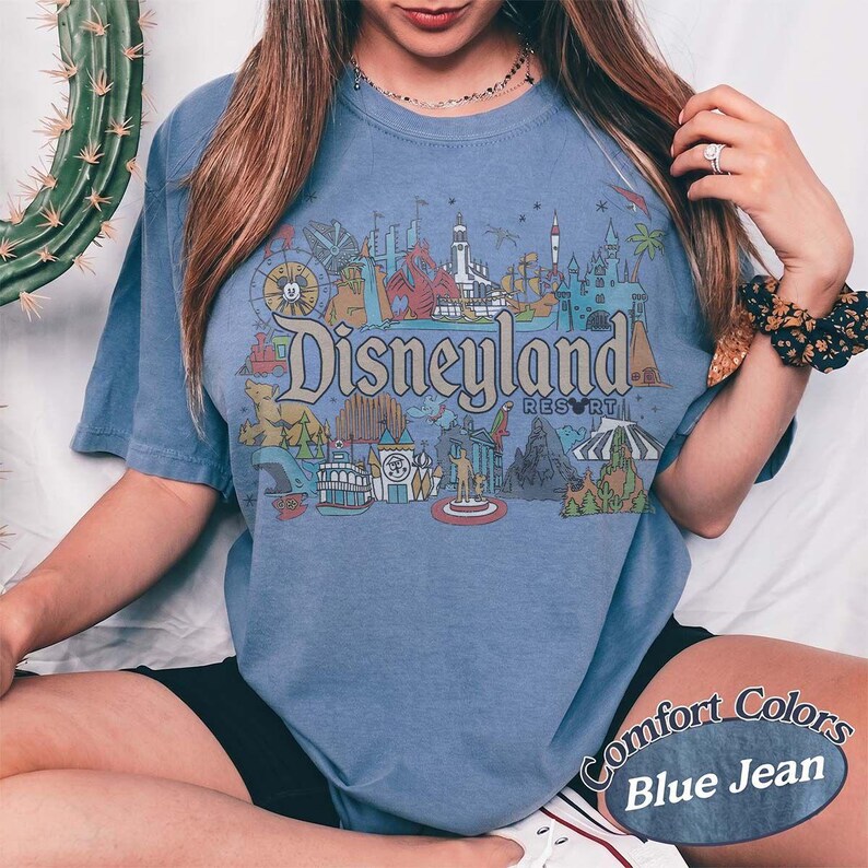 May include: Blue jean colored t-shirt featuring a Disneyland Resort graphic. The graphic includes various park icons in a colorful design. The shirt has short sleeves and a crew neck. The words "Comfort Colors" and "Blue Jean" are printed on the bottom right.