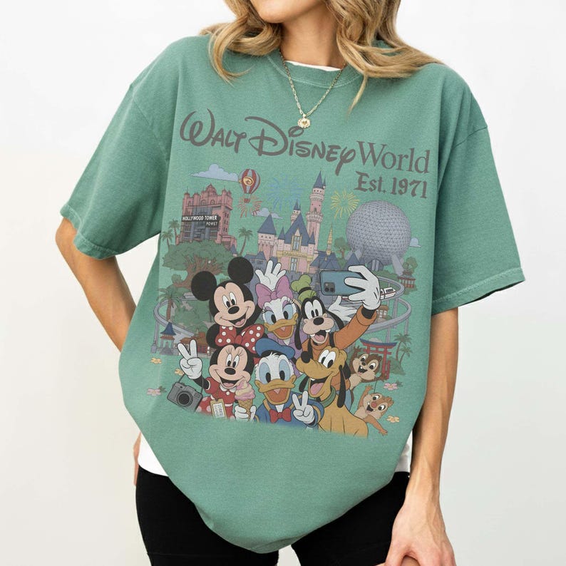 May include: A sage green short-sleeve t-shirt featuring a graphic print of Walt Disney World Est. 1971. The design includes Mickey Mouse, Minnie Mouse, Donald Duck, Daisy Duck, Goofy, Pluto, and Chip and Dale in front of park landmarks.