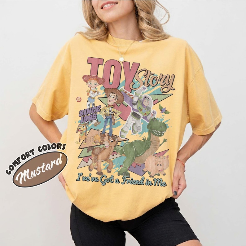 May include: A mustard yellow short-sleeve t-shirt featuring a vintage-style graphic of Toy Story characters including Woody, Buzz Lightyear, Jessie, Rex, Slinky Dog, and Hamm. The text reads Toy Story, Since 1995, and You've Got a Friend in Me.
