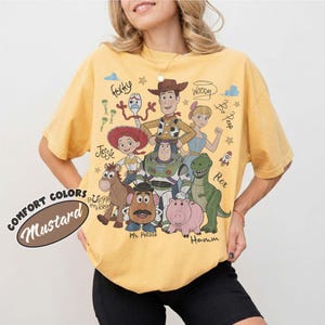 May include: A mustard yellow short-sleeve t-shirt featuring a graphic print of Toy Story characters including Woody, Buzz Lightyear, Jessie, Bullseye, Mr. Potato Head, Hamm, Bo Peep, and Forky, with their names written in a cursive script font.