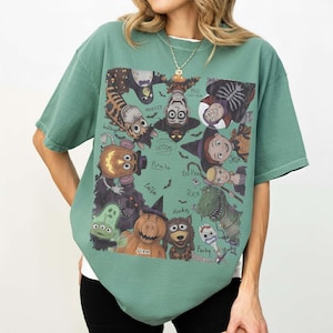May include: A sage green short-sleeve t-shirt featuring a rectangular graphic print of various cartoon characters dressed in Halloween costumes. The characters include a pumpkin-headed potato, a dinosaur, a pig, and others with handwritten labels.