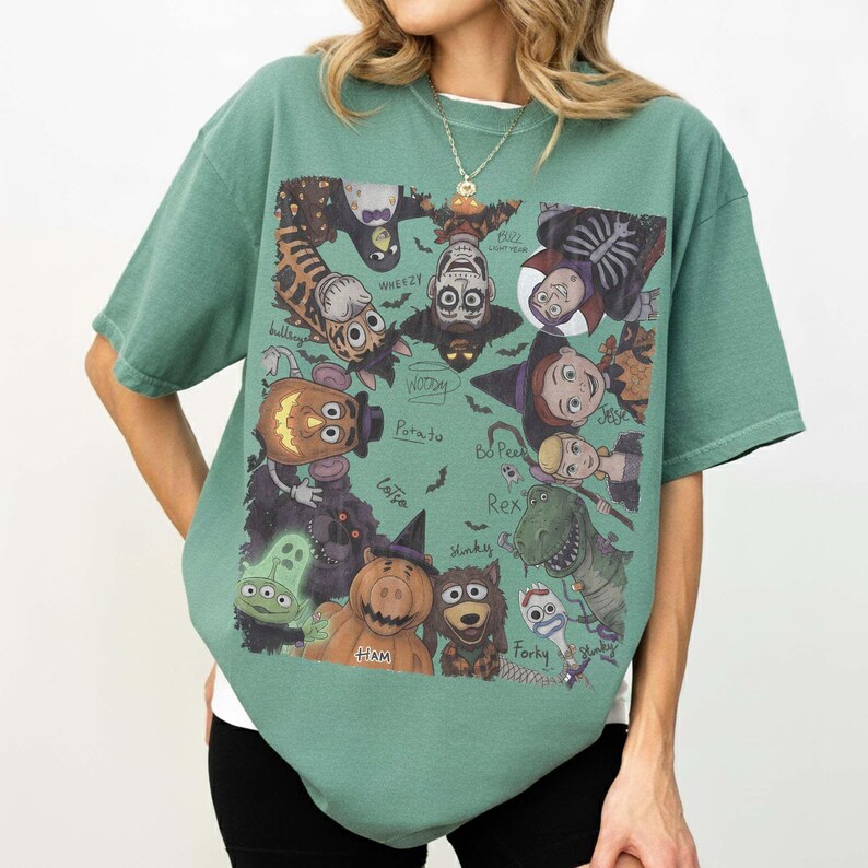 May include: A sage green short-sleeve t-shirt featuring a rectangular graphic print of various cartoon characters dressed in Halloween costumes. The characters include a pumpkin-headed potato, a dinosaur, a pig, and others with handwritten labels.