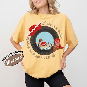 May include: A mustard yellow t-shirt featuring a graphic of a black tire framing a scene with cartoon characters. The design includes a red cowboy hat, a red cowboy boot, and the text Love has ways of bringing things back to life.