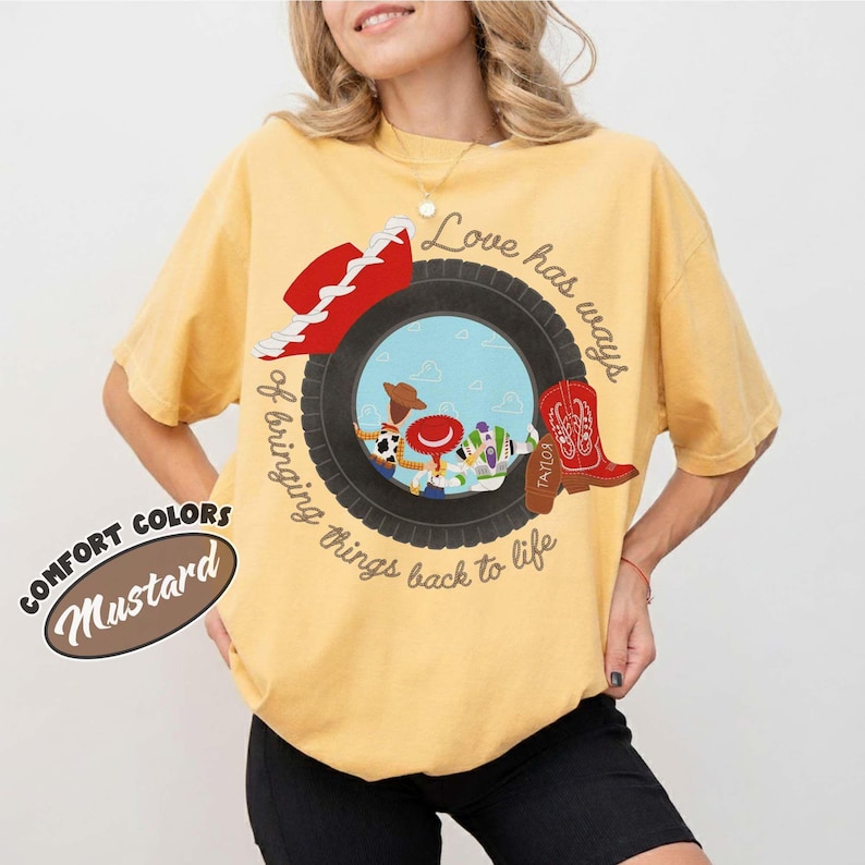 May include: A mustard yellow t-shirt featuring a graphic of a black tire framing a scene with cartoon characters. The design includes a red cowboy hat, a red cowboy boot, and the text Love has ways of bringing things back to life.