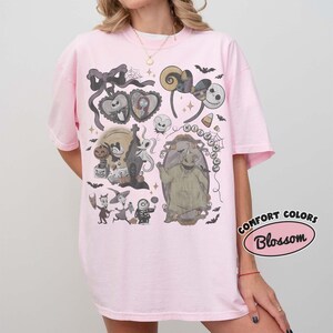 May include: A light pink t-shirt featuring a graphic print of characters and motifs from The Nightmare Before Christmas. The design includes Jack Skellington, Sally, Oogie Boogie, and various spooky elements like bats, spiderwebs, and a mirror.