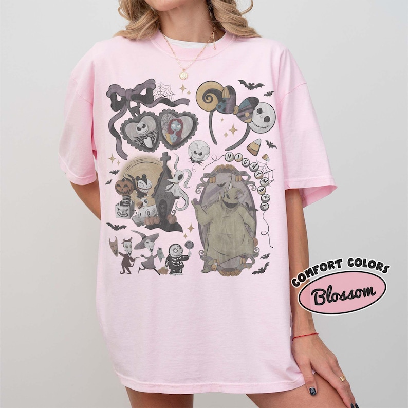 May include: A light pink t-shirt featuring a graphic print of characters and motifs from The Nightmare Before Christmas. The design includes Jack Skellington, Sally, Oogie Boogie, and various spooky elements like bats, spiderwebs, and a mirror.