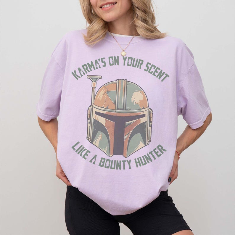 May include: A lilac t-shirt with a Mandalorian helmet graphic in brown, green, and orange. The text "Karma's on your scent like a bounty hunter" is printed above and below the helmet design, suitable for fans of the series.