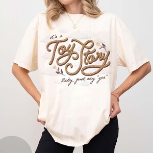 May include: An off-white short-sleeve t-shirt featuring a graphic design with the text "it's a Toy Story" written in a brown rope-like font, accompanied by the phrase "Baby, just say "yes"" and small illustrations of clouds and flowers.