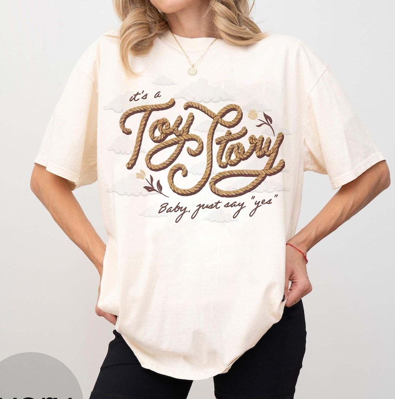 May include: An off-white short-sleeve t-shirt featuring a graphic design with the text "it's a Toy Story" written in a brown rope-like font, accompanied by the phrase "Baby, just say "yes"" and small illustrations of clouds and flowers.