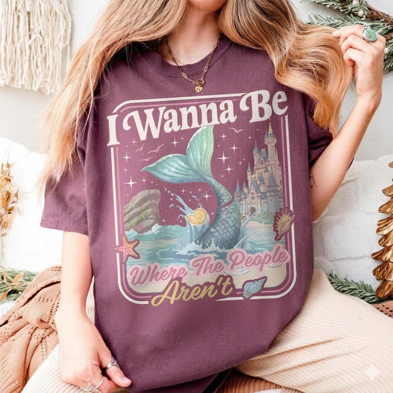 May include: Burgundy t-shirt featuring a mermaid illustration with a teal tail, a castle, and the text "I Wanna Be Where The People Aren't." The design includes a starfish and seashells. The shirt has short sleeves.