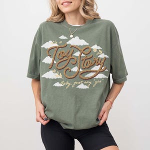 May include: An olive green short-sleeve t-shirt featuring a graphic of white clouds and the text "Toy Story" written in a brown rope-style font. Below the main text, the phrase "Baby, just say "yes"" is printed in a smaller, light yellow script.