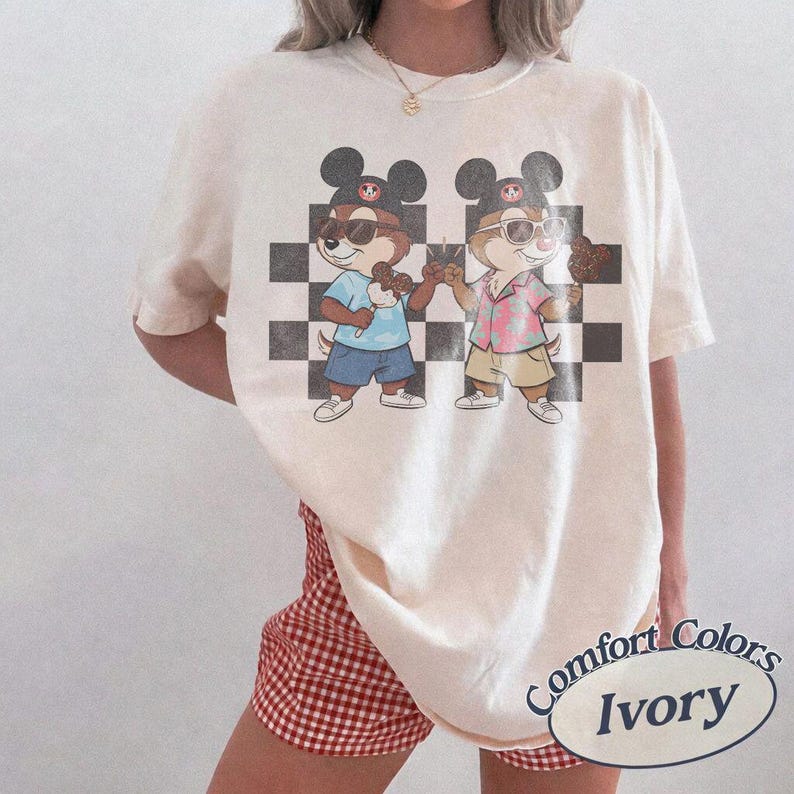 May include: Ivory-colored t-shirt featuring a graphic of two cartoon chipmunks wearing sunglasses and Mickey Mouse hats, holding ice cream cones. The chipmunks stand in front of a black and white checkered background. The shirt has the words "Comfort Colors Ivory" printed on it.