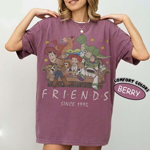 May include: A berry-colored short-sleeve t-shirt featuring a graphic print of Toy Story characters sitting on a couch with the text "FRIENDS SINCE 1995" below. The design includes Woody, Buzz Lightyear, Jessie, Rex, Bullseye, and Slinky Dog.