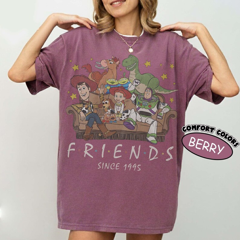 May include: A berry-colored short-sleeve t-shirt featuring a graphic print of Toy Story characters sitting on a couch with the text "FRIENDS SINCE 1995" below. The design includes Woody, Buzz Lightyear, Jessie, Rex, Bullseye, and Slinky Dog.