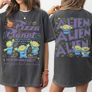 May include: Dark gray t-shirt featuring the Pizza Planet logo and alien characters. The front of the shirt has the text "Pizza Planet" and "Out of this world quality". The back of the shirt has the word "Alien" repeated three times.