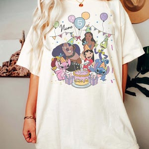 Pu&ograve; includere: T-shirt color avorio con una grafica colorata di personaggi dei cartoni animati che festeggiano un compleanno. Il design include una torta, palloncini e il numero 5. La parola "Name" &egrave; visibile nel design.