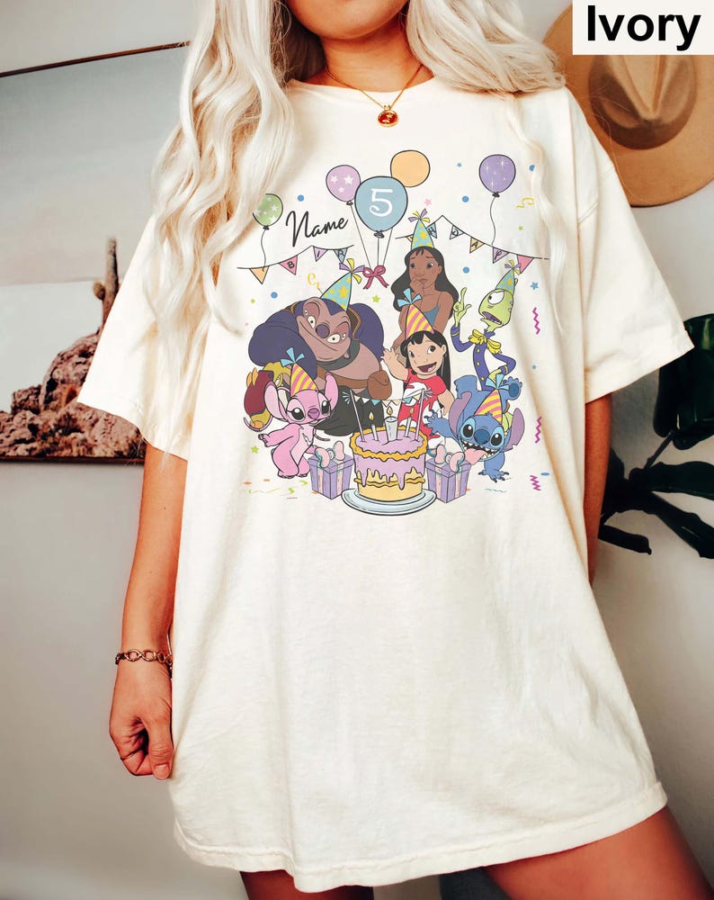 Pu&ograve; includere: T-shirt color avorio con una grafica colorata di personaggi dei cartoni animati che festeggiano un compleanno. Il design include una torta, palloncini e il numero 5. La parola "Name" &egrave; visibile nel design.