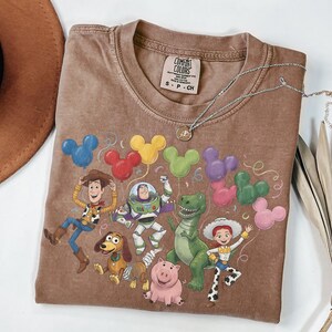 May include: A brown short-sleeve t-shirt featuring a graphic print of Toy Story characters including Woody, Buzz Lightyear, Rex, Slinky Dog, Jessie, and Hamm. The design includes colorful Mickey Mouse-shaped balloons and the Comfort Colors brand label.
