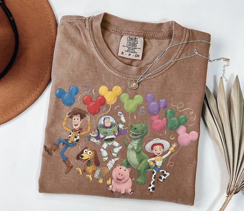 May include: A brown short-sleeve t-shirt featuring a graphic print of Toy Story characters including Woody, Buzz Lightyear, Rex, Slinky Dog, Jessie, and Hamm. The design includes colorful Mickey Mouse-shaped balloons and the Comfort Colors brand label.