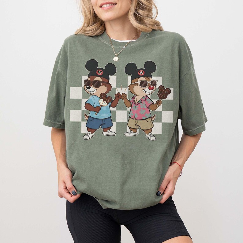 May include: Green t-shirt featuring a cartoon illustration of two chipmunks wearing sunglasses and Mickey Mouse hats. One chipmunk holds an ice cream cone, the other a chocolate-covered ice cream bar. The background has a checkered pattern.