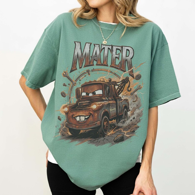 Puede incluir: Camiseta verde de manga corta con un gr&aacute;fico de Mate de la pel&iacute;cula Cars. La gr&uacute;a marr&oacute;n tiene una cara sonriente, rodeada de pernos y la palabra "MATER" en una fuente desgastada.