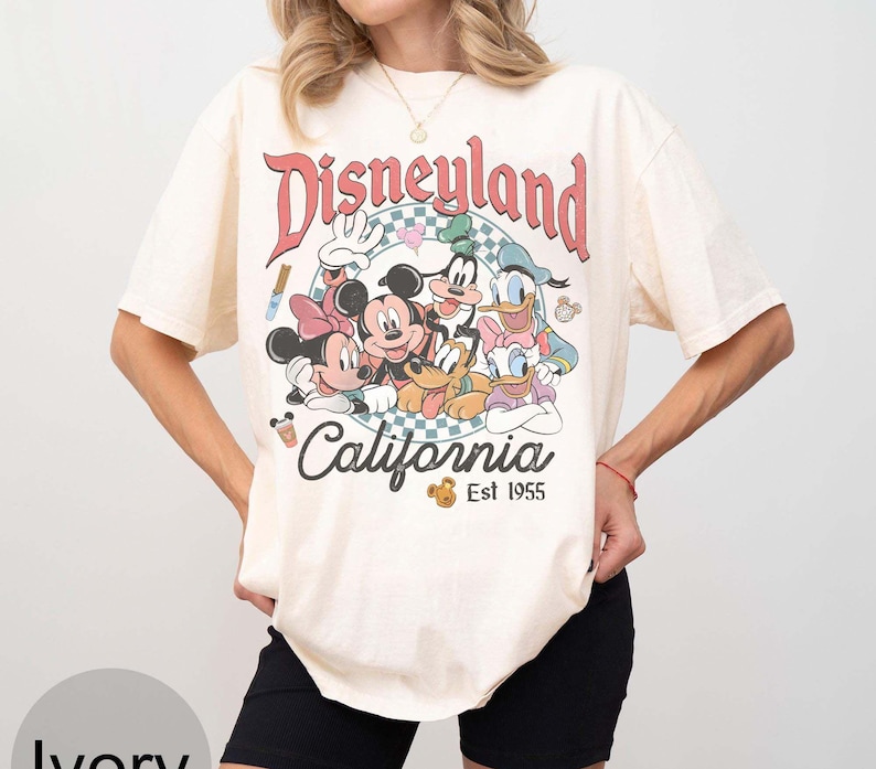 May include: Ivory-colored t-shirt featuring a Disneyland graphic with Mickey Mouse, Minnie Mouse, Donald Duck, Daisy Duck, Goofy, and Pluto. The text "Disneyland California Est 1955" is printed above and below the characters.