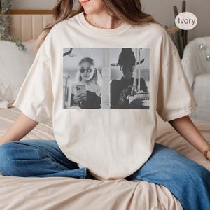 May include: Ivory-colored t-shirt featuring a black and white image of two people. One person is wearing sunglasses and holding a phone, while the other is wearing a hat. The shirt is a relaxed fit and has a crew neck.