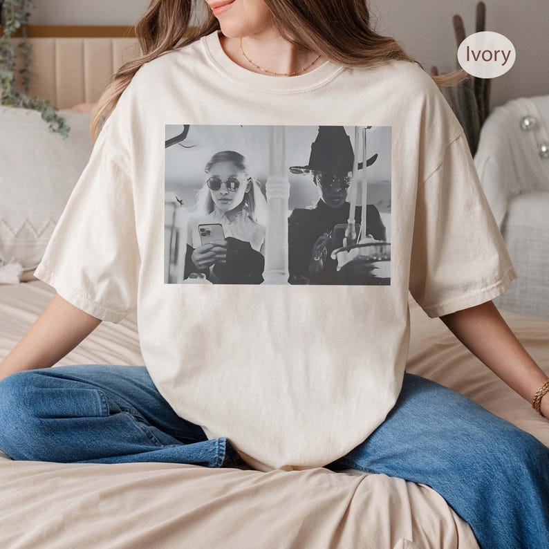 May include: Ivory-colored t-shirt featuring a black and white image of two people. One person is wearing sunglasses and holding a phone, while the other is wearing a hat. The shirt is a relaxed fit and has a crew neck.