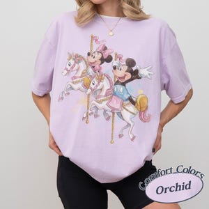 Op de afbeelding: Een lichtpaars T-shirt met een carrouselontwerp met Mickey en Minnie Mouse op witte paarden. Het shirt heeft een losse pasvorm en de woorden "Comfort Colors Orchid" staan rechtsonder.