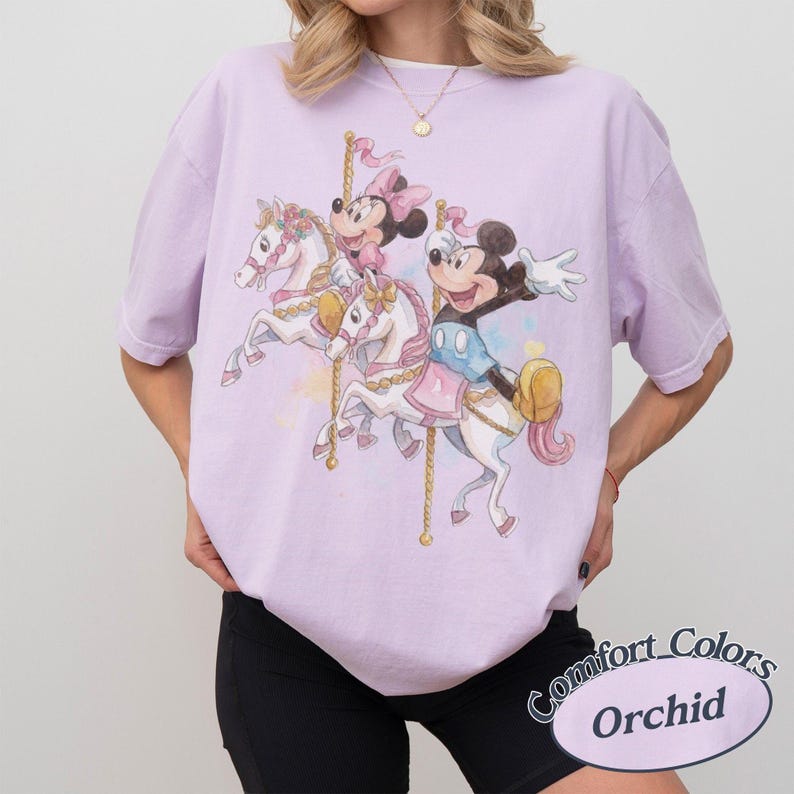 Op de afbeelding: Een lichtpaars T-shirt met een carrouselontwerp met Mickey en Minnie Mouse op witte paarden. Het shirt heeft een losse pasvorm en de woorden "Comfort Colors Orchid" staan rechtsonder.