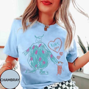 May include: Light blue t-shirt featuring a whimsical drawing of a monster with pink polka dots, a heart that says "Kitty Boo," and a small figure. The design is in a crayon-like style.
