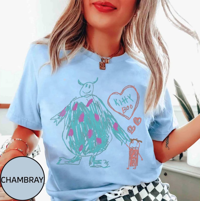 May include: Light blue t-shirt featuring a whimsical drawing of a monster with pink polka dots, a heart that says "Kitty Boo," and a small figure. The design is in a crayon-like style.