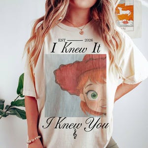 May include: A cream-colored short-sleeve t-shirt featuring a graphic of a cartoon character with red hair and a red hat. The text reads EST 2026 I Knew It I Knew You with a small musical note symbol below the text, printed in a black serif font.