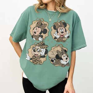 May include: Green t-shirt featuring four Minnie Mouse illustrations in leopard print outfits. Each illustration is set within a decorative frame. The shirt has short sleeves and a relaxed fit.