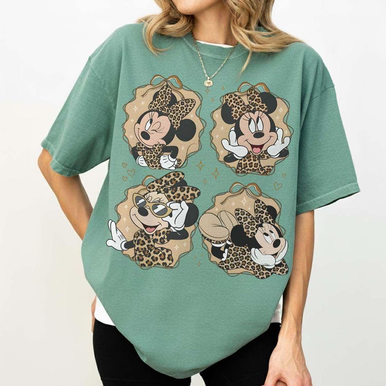 May include: Green t-shirt featuring four Minnie Mouse illustrations in leopard print outfits. Each illustration is set within a decorative frame. The shirt has short sleeves and a relaxed fit.
