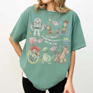 K&ouml;nnte beinhalten: Gr&uuml;nes T-Shirt mit Figuren aus dem Film Toy Story, darunter Buzz Lightyear, Woody, Jessie, Rex, Hamm, Slinky Dog, Bullseye und die kleinen gr&uuml;nen M&auml;nnchen. Der Text "You've Got a Friend in Me" ist ebenfalls enthalten.