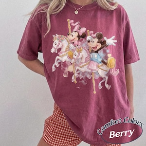 Op de afbeelding: Bessenkleurig T-shirt met een carrouselontwerp met Mickey en Minnie Mouse-personages. De afbeelding bevat witte paarden en pastelkleuren. Het shirt heeft korte mouwen en een relaxte pasvorm. De tekst "Comfort Colors Berry" staat rechtsonder.