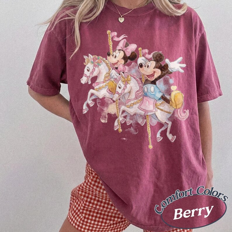 Op de afbeelding: Bessenkleurig T-shirt met een carrouselontwerp met Mickey en Minnie Mouse-personages. De afbeelding bevat witte paarden en pastelkleuren. Het shirt heeft korte mouwen en een relaxte pasvorm. De tekst "Comfort Colors Berry" staat rechtsonder.
