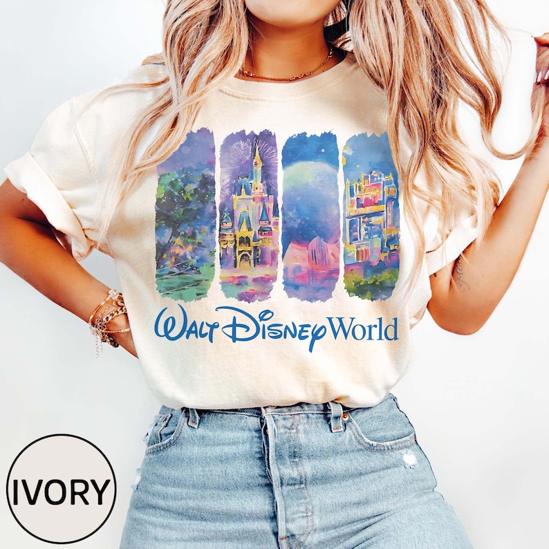 May include: Ivory-colored t-shirt featuring a colorful graphic of Walt Disney World landmarks. The design includes illustrations of a castle, fireworks, and other park features. The words "Walt Disney World" are printed in blue.