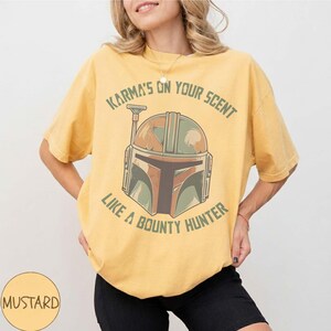 May include: Mustard-coloured t-shirt with a bounty hunter helmet graphic and the text "Karma's on your scent like a bounty hunter." The design uses green, brown, and orange. The shirt has a relaxed fit and is a casual wear item.