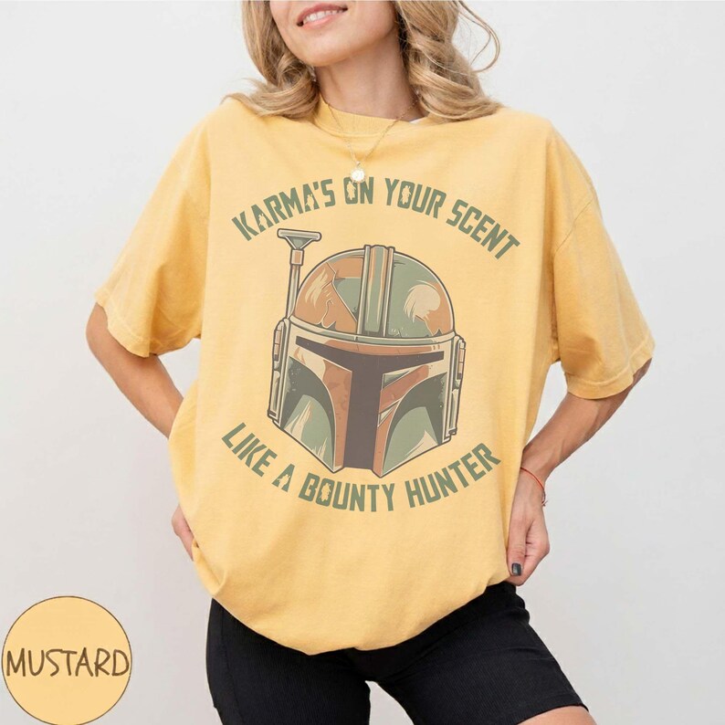 May include: Mustard-coloured t-shirt with a bounty hunter helmet graphic and the text "Karma's on your scent like a bounty hunter." The design uses green, brown, and orange. The shirt has a relaxed fit and is a casual wear item.