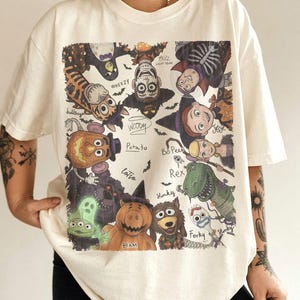 May include: An off-white t-shirt featuring a graphic print of various animated characters dressed in Halloween costumes. The characters include Woody, Buzz Lightyear, Jessie, Bo Peep, Rex, Forky, Ham, and others, with their names written in cursive script.