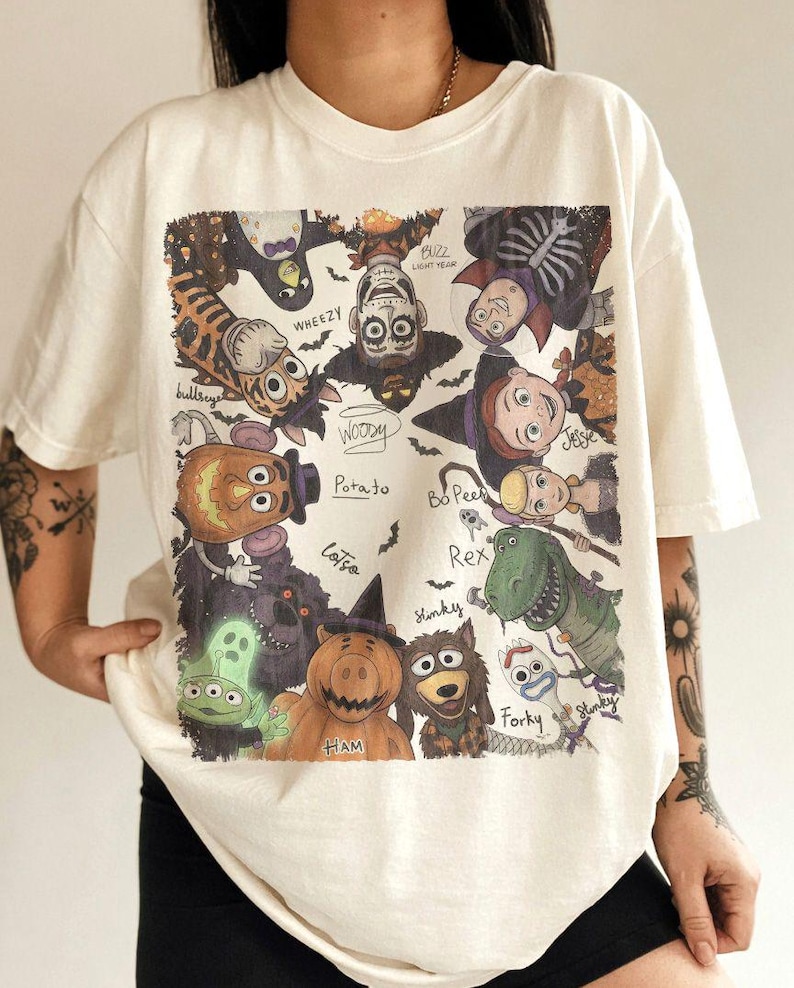 May include: An off-white t-shirt featuring a graphic print of various animated characters dressed in Halloween costumes. The characters include Woody, Buzz Lightyear, Jessie, Bo Peep, Rex, Forky, Ham, and others, with their names written in cursive script.