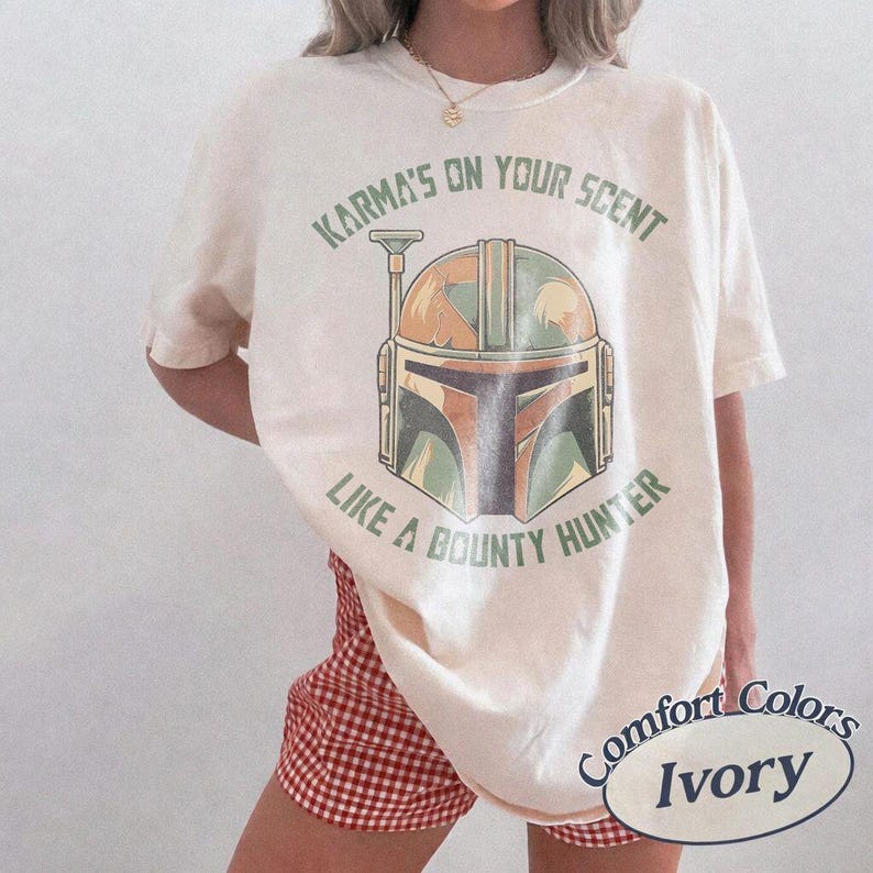 May include: Cream-coloured t-shirt featuring a helmet graphic and the text "Karma's on your scent like a bounty hunter." The shirt is a Comfort Colors brand. The model is wearing red and white checkered shorts.