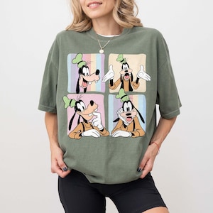 Może przedstawiać: Oliwkowo-zielona koszulka z czteropanelową grafiką Goofy'ego w r&oacute;żnych pozach. Każdy panel ma inne pastelowe tło. Goofy ma na sobie charakterystyczny zielony kapelusz i pomarańczową koszulę. Koszulka jest w stylu casual.
