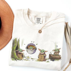 May include: A cream-colored short-sleeve t-shirt featuring a graphic print of four small green creatures in various poses, including one in a floating pod. The shirt has a Comfort Colors brand label at the neck and a crew neckline design.