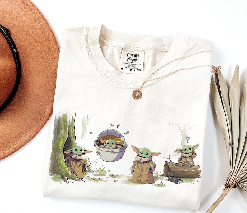 May include: A cream-colored short-sleeve t-shirt featuring a graphic print of four small green creatures in various poses, including one in a floating pod. The shirt has a Comfort Colors brand label at the neck and a crew neckline design.