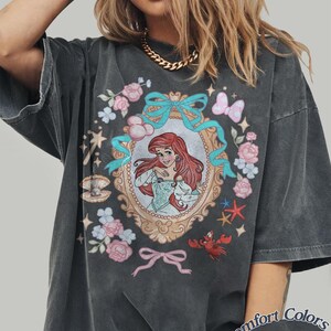 Puede incluir: Camiseta gris oscuro Comfort Colors Pepper con una ilustraci&oacute;n enmarcada de una sirena pelirroja rodeada de flores, lazos y criaturas marinas. La camiseta tiene un aspecto vintage y desgastado.