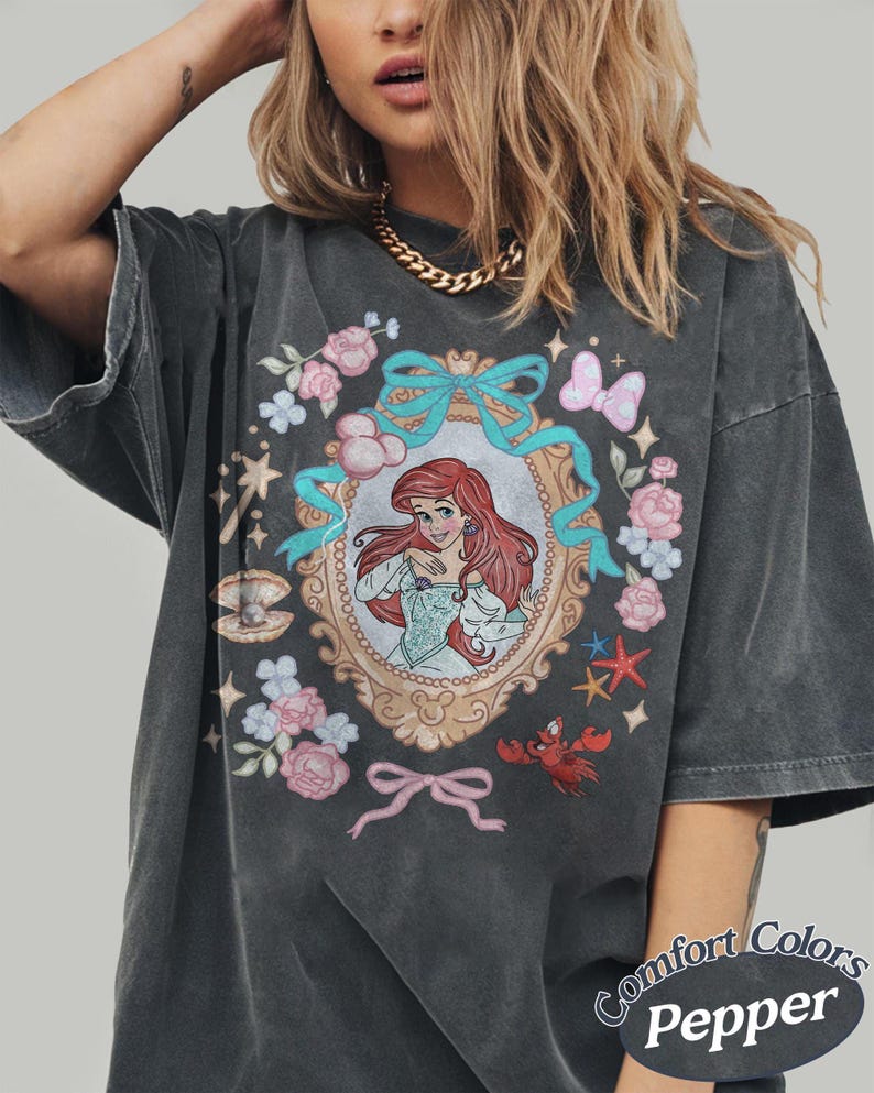 Puede incluir: Camiseta gris oscuro Comfort Colors Pepper con una ilustraci&oacute;n enmarcada de una sirena pelirroja rodeada de flores, lazos y criaturas marinas. La camiseta tiene un aspecto vintage y desgastado.