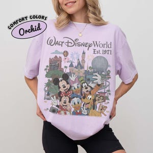 May include: An orchid-colored t-shirt featuring a graphic print of Walt Disney World Est. 1971. The design includes illustrations of Mickey Mouse, Minnie Mouse, Donald Duck, Daisy Duck, Goofy, Pluto, Chip, and Dale in front of park landmarks.